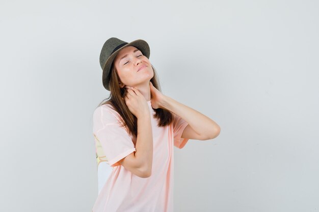 Jonge vrouw die lijdt aan nekpijn in roze t-shirt, hoed en op zoek naar ongemakkelijk, vooraanzicht.