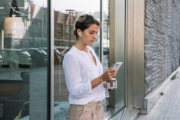 Jonge vrouw die meeneemkoffiekop houdt die mobiele telefoon met behulp van