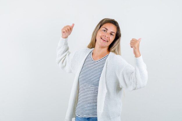 Jonge vrouw die met duimen in t-shirt, jasje terug richt en gelukkig kijkt. vooraanzicht.