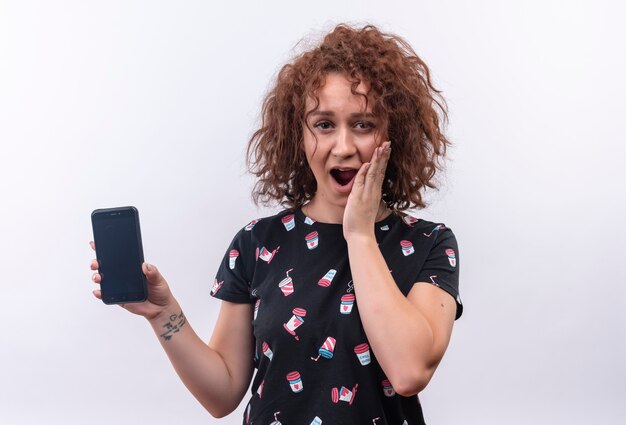 Jonge vrouw die met kort krullend haar smartphone houdt die verbaasd en verbaasd over witte muur kijkt