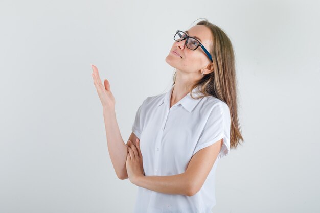 Jonge vrouw die met opgeheven palm in wit t-shirt omhoog kijkt en hoopvol kijkt.