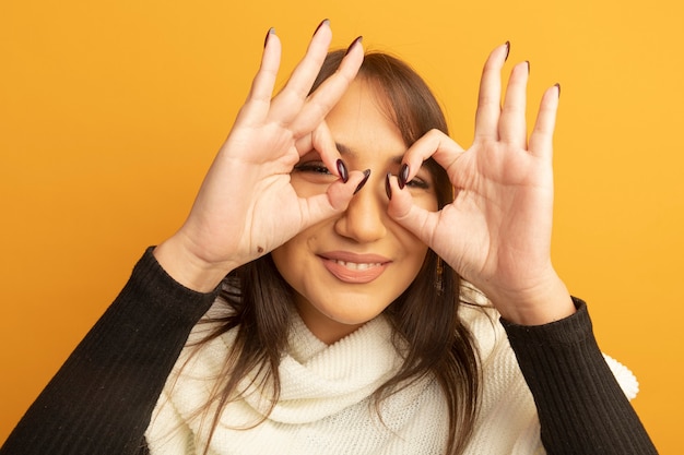 Gratis foto jonge vrouw die met witte sjaal ok teken als verrekijker gebaar maakt die door vingers vrolijk glimlachen kijken