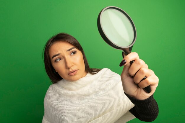 Jonge vrouw die met witte sjaal vergrootglas met ernstig gezicht bekijkt