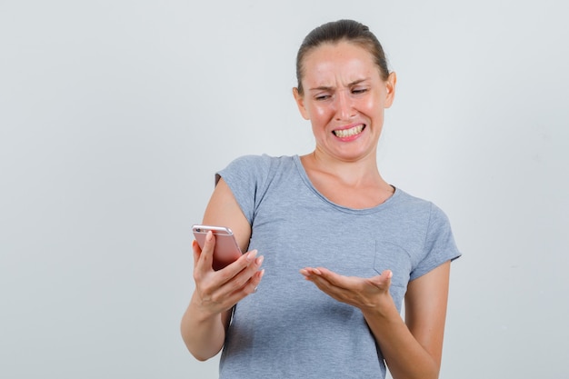 Jonge vrouw die mobiele telefoon in grijs t-shirt bekijkt en geïrriteerd kijkt. vooraanzicht.