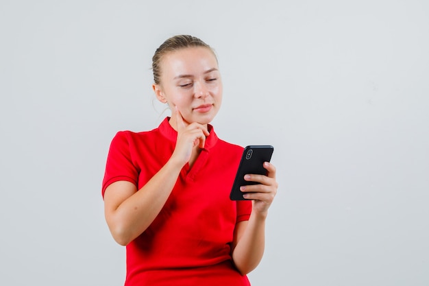 Jonge vrouw die mobiele telefoon in rood t-shirt bekijkt en peinzend kijkt