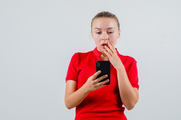 Jonge vrouw die mobiele telefoon in rood t-shirt bekijkt en verbaasd kijkt