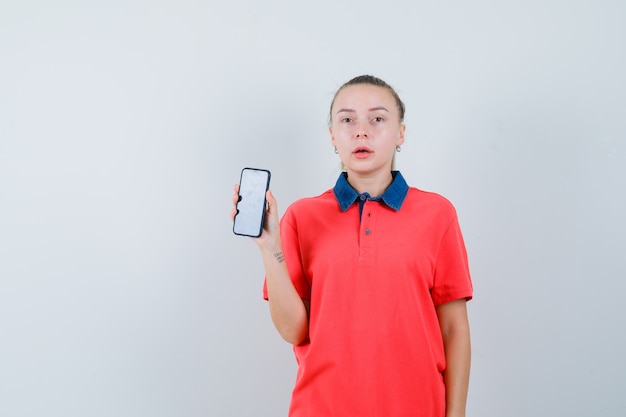 Jonge vrouw die mobiele telefoon in t-shirt toont en verbaasd kijkt