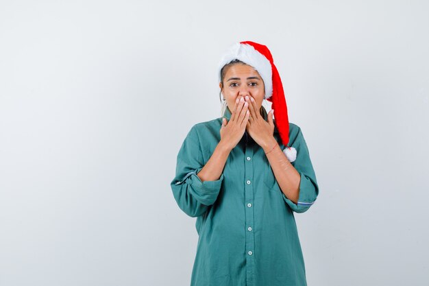 Jonge vrouw die mond bedekt met handen in shirt, kerstmuts en verbaasd kijkt, vooraanzicht.