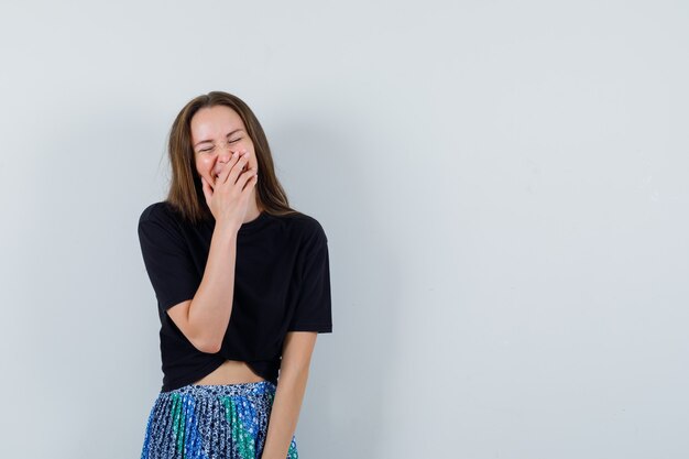 Jonge vrouw die mond behandelt met handen en in zwart t-shirt en blauwe rok lacht en aantrekkelijk kijkt