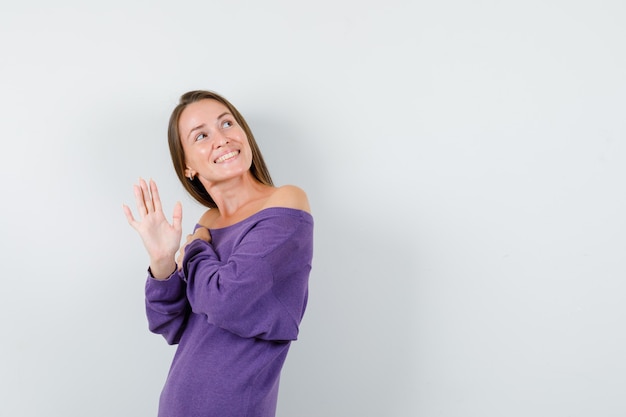 Jonge vrouw die naar boven kijkt en zwaait hand in violet overhemd en op zoek vrolijk, vooraanzicht.