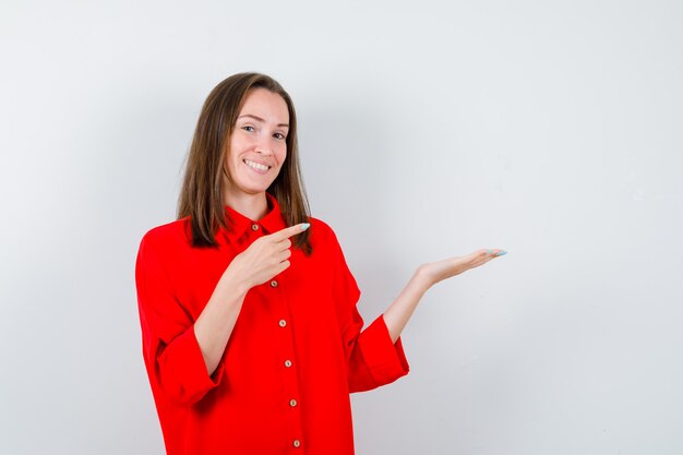 Jonge vrouw die naar haar handpalm wijst in een rode blouse opzij en er vrolijk uitziet, vooraanzicht.