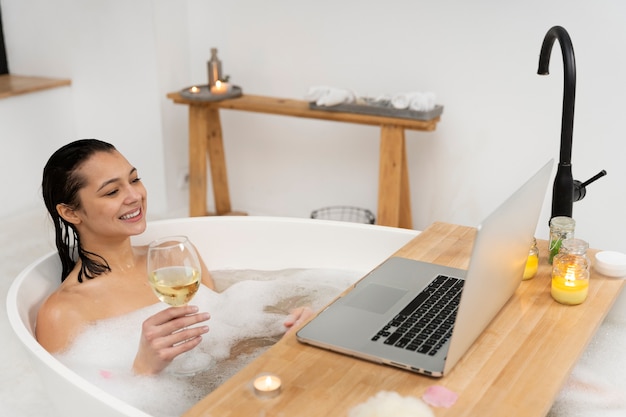 Jonge vrouw die naar haar laptop kijkt en wijn drinkt terwijl ze een bad neemt
