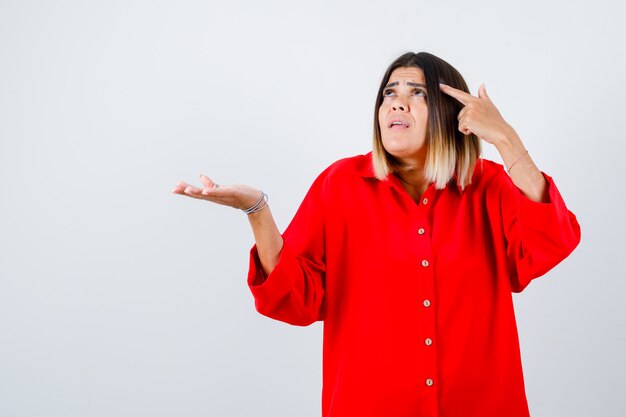 Jonge vrouw die naar het hoofd wijst terwijl ze iets in een rood oversized shirt vasthoudt en aarzelend kijkt, vooraanzicht.