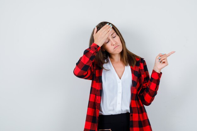 Jonge vrouw die naar rechts wijst, met de hand op het voorhoofd in vrijetijdskleding en er vermoeid uitziet. vooraanzicht.