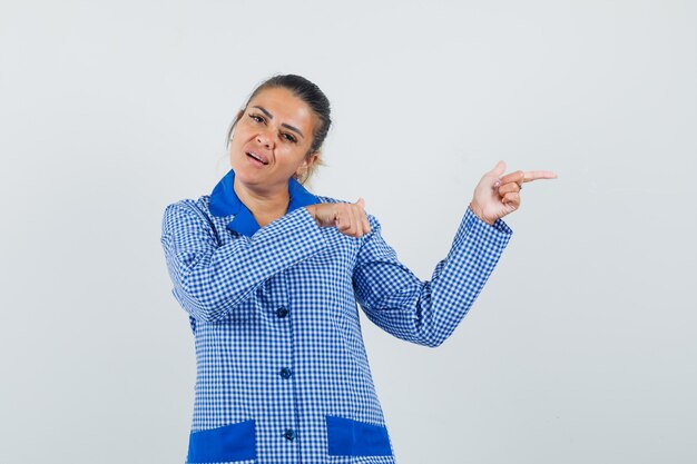 Jonge vrouw die naar rechts wijst met wijsvingers in blauw geruit pyjama-shirt en er mooi uitziet, vooraanzicht.