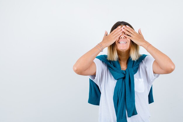 Jonge vrouw die ogen bedekt met handen in wit t-shirt en er gelukkig uitziet, vooraanzicht.