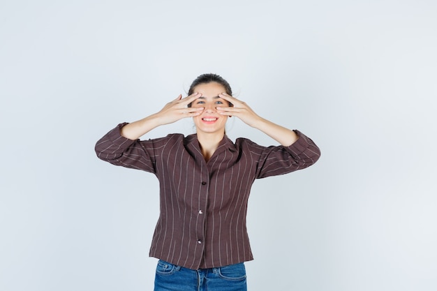 Jonge vrouw die ogen opent met vingers, glimlachend in gestreept shirt, spijkerbroek en vrolijk, vooraanzicht.