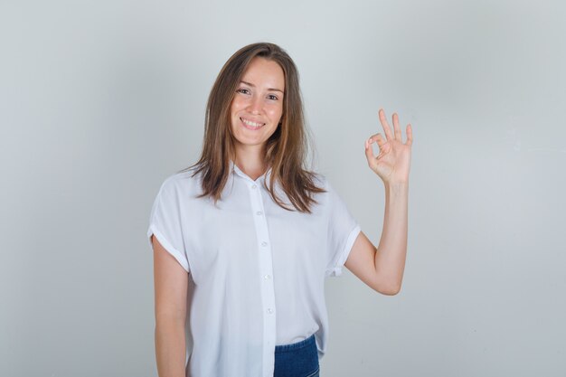 Jonge vrouw die ok gebaar in wit t-shirt, jeans toont en vrolijk kijkt