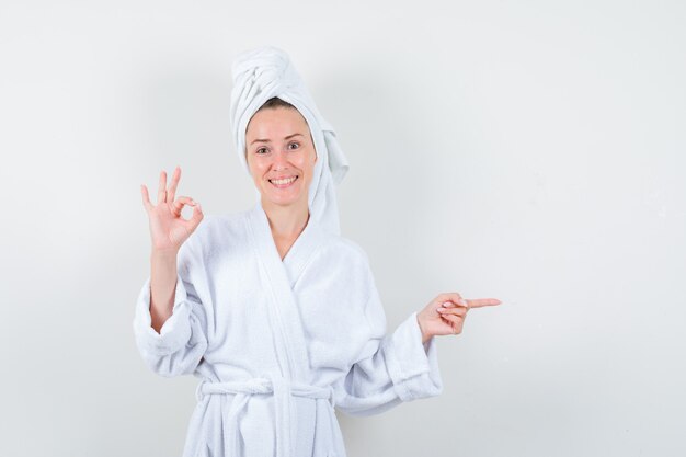 Jonge vrouw die ok gebaar toont, naar rechts wijst in witte badjas, handdoek en er vrolijk uitziet, vooraanzicht.