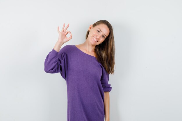 Jonge vrouw die ok teken in violet overhemd toont en gelukkig, vooraanzicht kijkt.