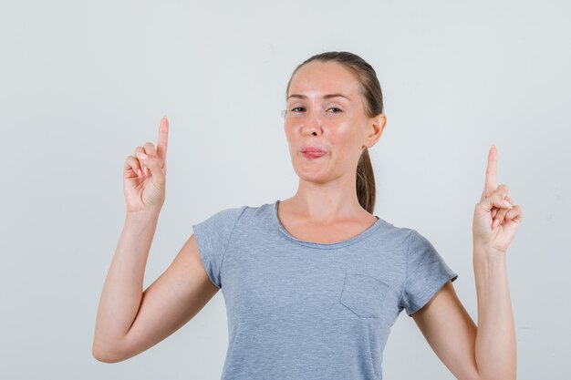 Jonge vrouw die omhoog wijst en tong in grijs t-shirt, glazen vooraanzicht toont.