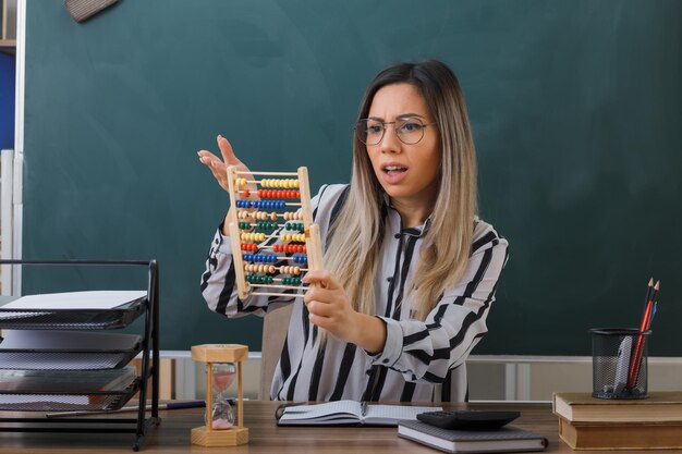 jonge vrouw die onderwijzers zit aan de schoolbank voor het bord in de klas en uitleg geeft over de les met het telraam dat er verward en ontevreden uitziet