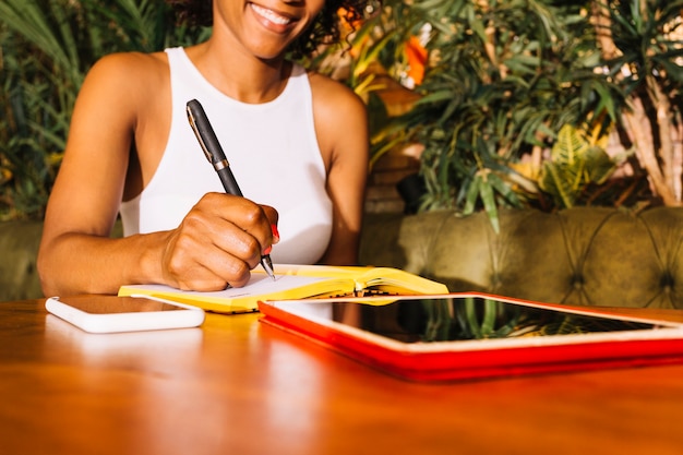 Jonge vrouw die op agenda met pen over de houten lijst met cellphone en digitale tablet schrijft