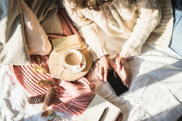 Jonge vrouw die op bed met kop van koffie ligt en smartphone gebruikt