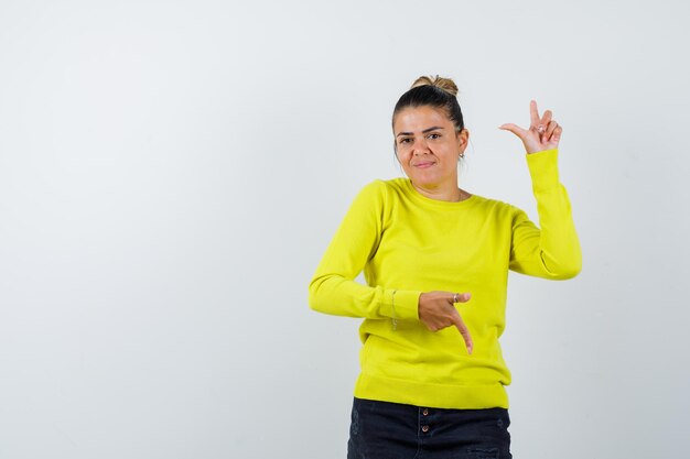 Jonge vrouw die op en neer wijst in gele trui en zwarte broek en er gelukkig uitziet