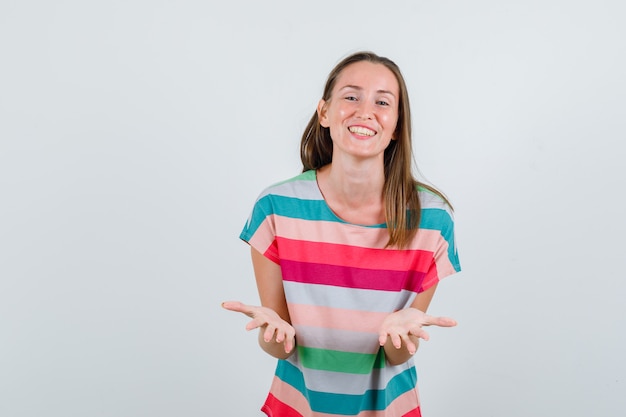 Jonge vrouw die palmen in vragend gebaar in t-shirt opheft en positief, vooraanzicht kijkt.