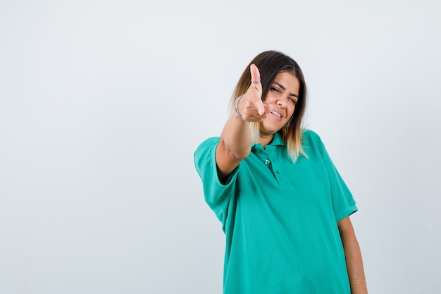 Jonge vrouw die pistoolgebaar naar camera in polot-shirt toont en er vrolijk uitziet, vooraanzicht.