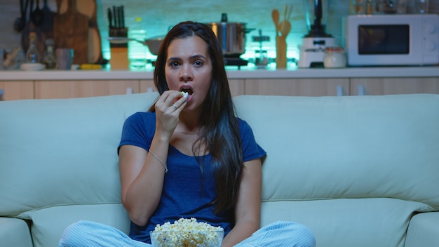 Jonge vrouw die popcorn eet en naar een interessante serie op tv kijkt. Geschokt geconcentreerd verbaasd alleen thuis 's nachts dame met verrast gezicht kijkend naar een spannende film zittend op een comfortabele bank