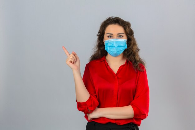 Jonge vrouw die rode blouse in medisch beschermend masker draagt die camera met ernstig gezicht bekijkt dat met vinger aan de kant over geïsoleerde witte achtergrond richt