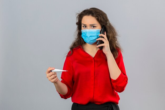 Jonge vrouw die rode blouse in medisch beschermend masker draagt die op mobiele telefoon spreekt die bezorgd kijkt die digitale thermometer over geïsoleerde witte achtergrond houdt