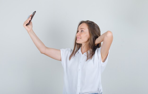 Jonge vrouw die selfie met hand achter hoofd in t-shirt neemt en vrolijk kijkt
