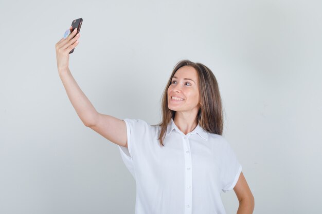 Jonge vrouw die selfie op telefoon in t-shirt neemt en vrolijk kijkt