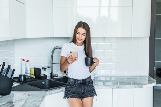 Jonge vrouw die smartphone gebruiken die bij keukenlijst leunen met koffiemok in een modern huis. Het glimlachen van de telefoonbericht van de vrouwenlezing. Donkerbruin gelukkig meisje die een tekstbericht typen