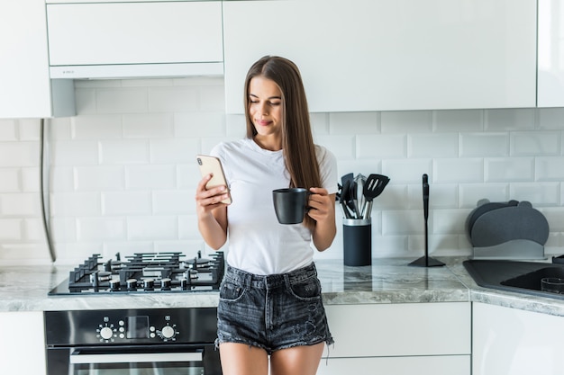 Jonge vrouw die smartphone gebruiken die bij keukenlijst leunen met koffiemok in een modern huis. Het glimlachen van de telefoonbericht van de vrouwenlezing. Donkerbruin gelukkig meisje die een tekstbericht typen