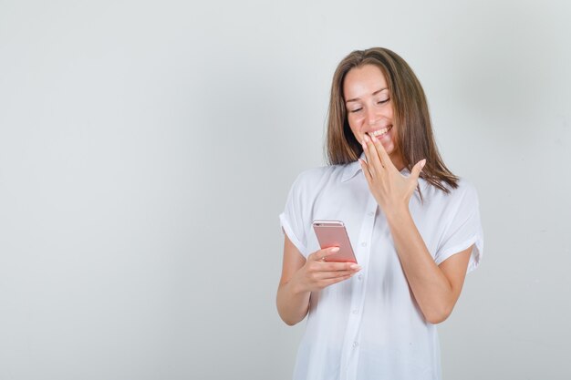 Jonge vrouw die smartphone met hand op mond in wit t-shirt gebruikt en gelukkig kijkt