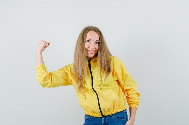 Jonge vrouw die spieren in geel bomberjack en blauwe jean toont en zelfverzekerd, vooraanzicht kijkt.