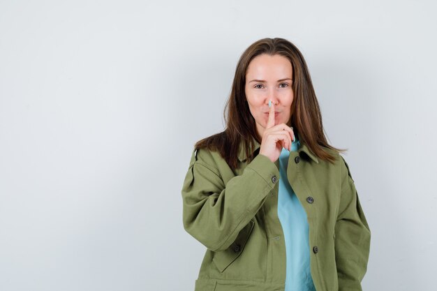 Jonge vrouw die stiltegebaar in t-shirt, jasje toont en vrolijk kijkt. vooraanzicht.