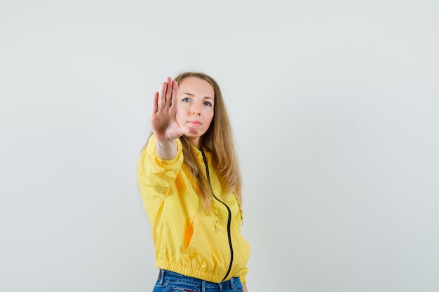 Jonge vrouw die stopbord in geel bomberjack en blauwe jean toont en ernstig, vooraanzicht kijkt.