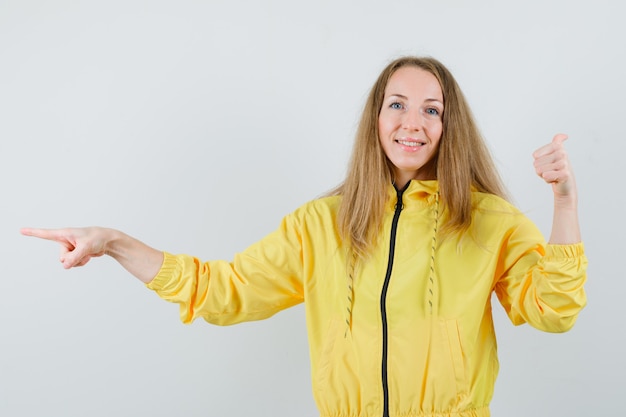 Jonge vrouw die tegengestelde richtingen in gele bomberjack en blauwe jean richt en optimistisch, vooraanzicht kijkt.