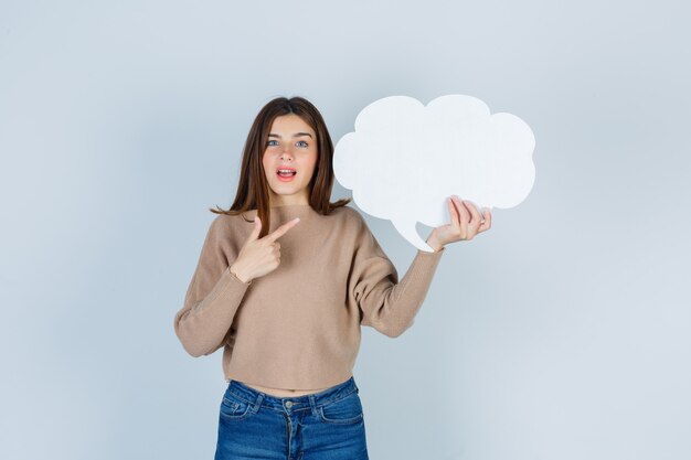 Jonge vrouw die tekstballon in trui, jeans toont en verbaasd kijkt, vooraanzicht.