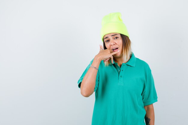 Jonge vrouw die telefoongebaar maakt in polot-shirt, muts en perplex kijkt. vooraanzicht.