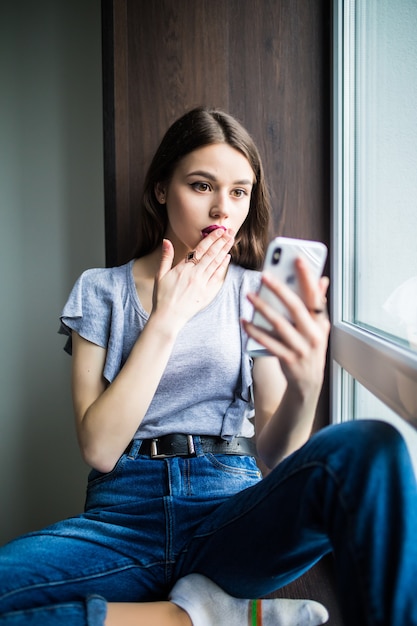 Jonge vrouw die thuis op een vensterbank zit en sms't op haar telefoon communicatie vrouwelijk uitziend bericht