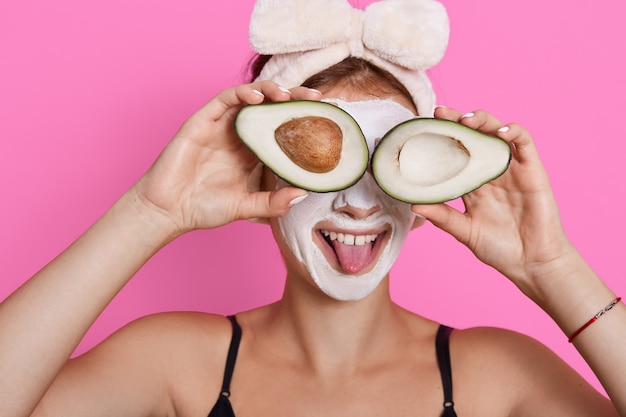 Jonge vrouw die twee helften avocado vasthoudt en de ogen ermee bedekt, haar tong laat zien, een haarband draagt, een vochtinbrengend masker op haar gezicht heeft, plezier heeft terwijl ze thuis schoonheidsprocedures doet.