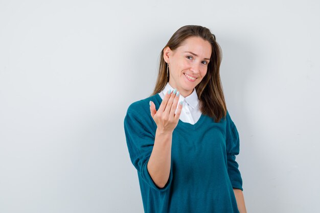 Jonge vrouw die uitnodigt om in een trui over een wit overhemd te komen en er vrolijk uitziet. vooraanzicht.