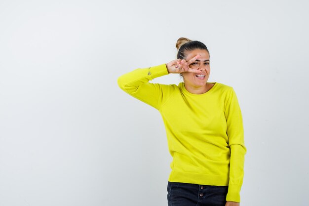 Jonge vrouw die v-teken op oog in gele sweater en zwarte broek toont en gelukkig kijkt
