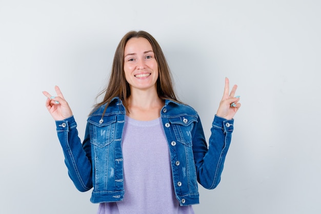 Jonge vrouw die vredesgebaar in t-shirt, jas toont en er vrolijk uitziet, vooraanzicht.